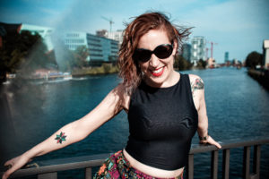 A photo of a smiling Jillian York, wearing sunglasses and showing off her tattoos while standing on a Berlin bridge